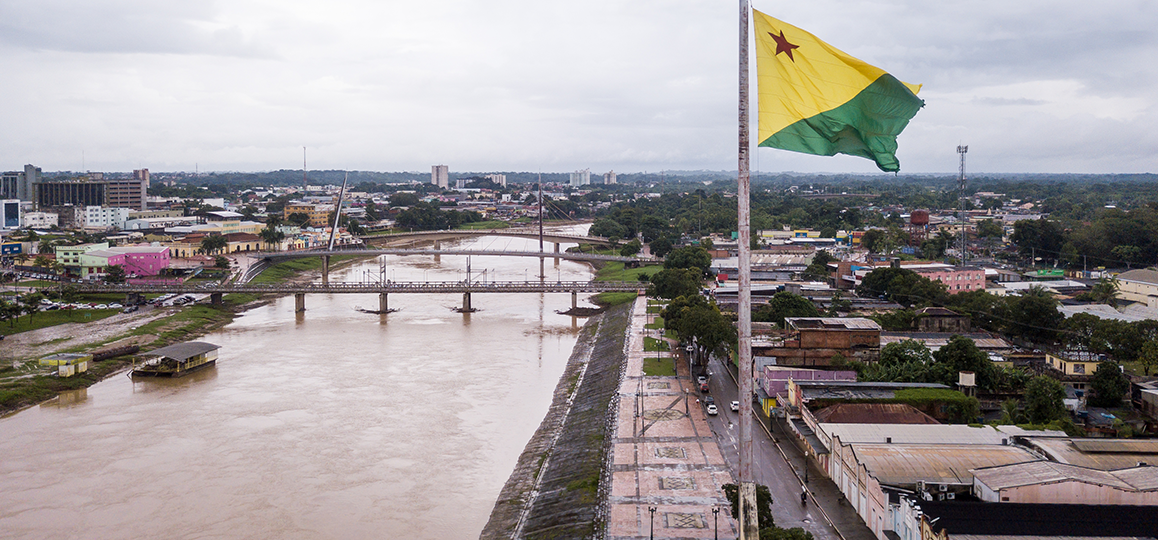 Foto de um rio que corta a cidade de Rio Brando (AC), com edifícios e casas em suas duas margens, conectadas por uma ponte. O céu está nebuloso e a bandeira do Acre em primeiro plano.