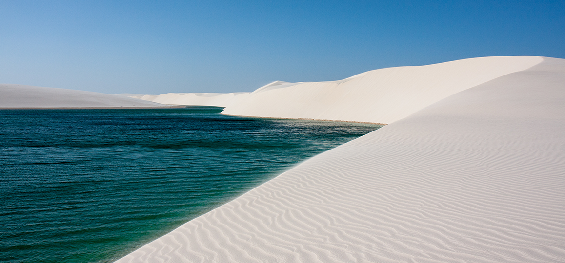 Foto de dunas de areia branca que contornam uma piscina de água nos Lençóis Maranhenses (MA).