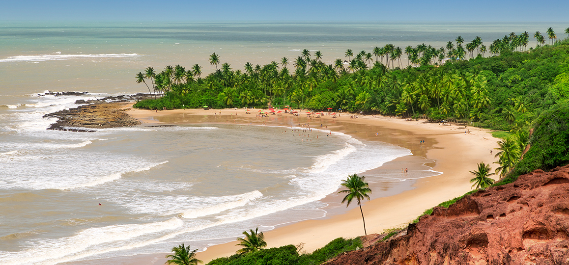 Foto da vista da praia do Coqueirinho, em João Pessoa (PB), com uma rocha em primeiro plano, em uma das pontas da praia, e coqueiral em segundo plano, na outra ponta da praia.