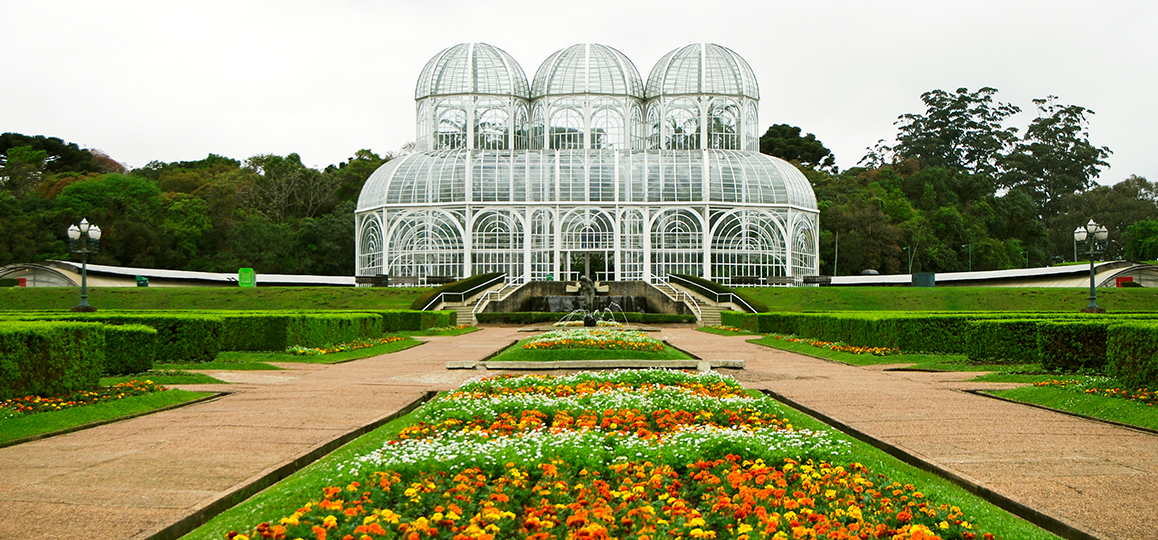 Foto de um jardim e, ao fundo, está a estrutura de metal da estufa do Jardim Botânico de Curitiba