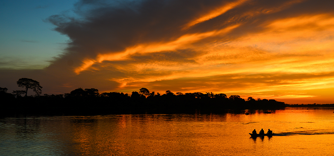 Foto do rio Guaporé Dourado (RO) ao entardecer, com luz natural dourada refletida nas nuvens e na água, e a silhueta de um pequeno barco com três pessoas cruzando o rio.