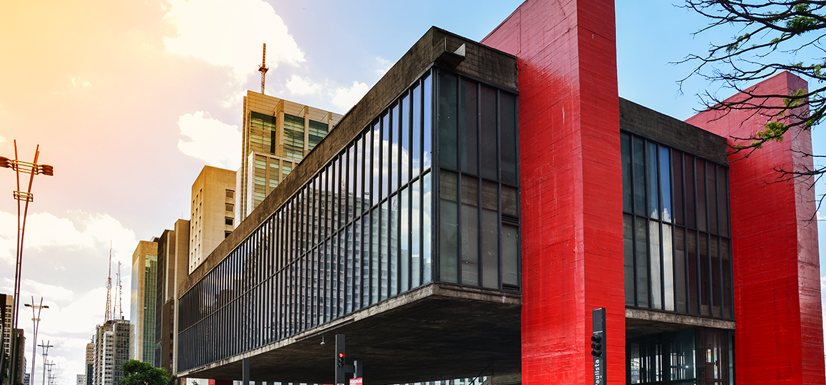 Foto do prédio modernista do MASP, em São Paulo (SP) - estrutura retangular com janelas de vidro sustentada por vigas vermelhas que contornam as laterais e o topo do prédio.