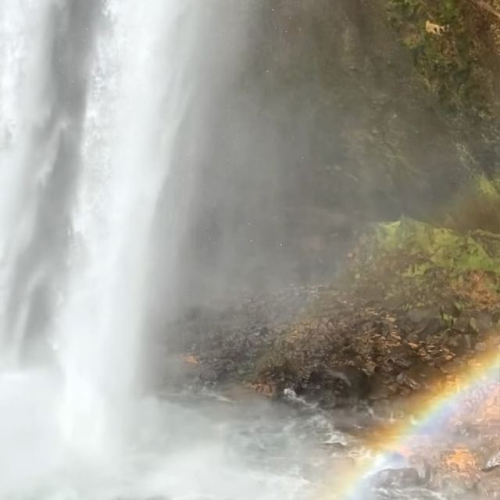 Vídeo de uma mulher sentada em uma pedra, na beira de uma cachoeira. Na paisagem, há um arco-íris formado pela queda d'água