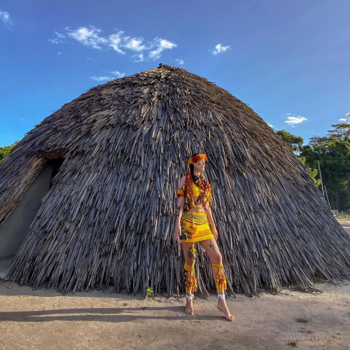 Foto de uma mulher em pé, em frente a uma oca, usando trajes tradicionais de uma comunidade indígena.